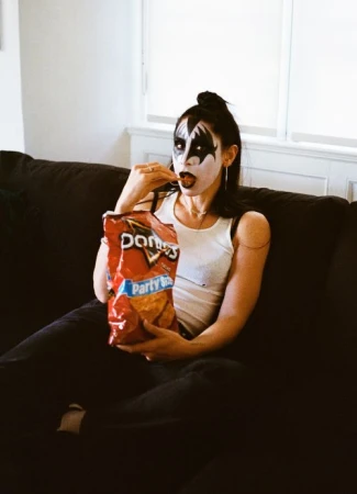 Woman with KISS-style makeup sits on a couch while eating Doritos; white walls, black hair, white tank top, and a red Doritos bag visible.