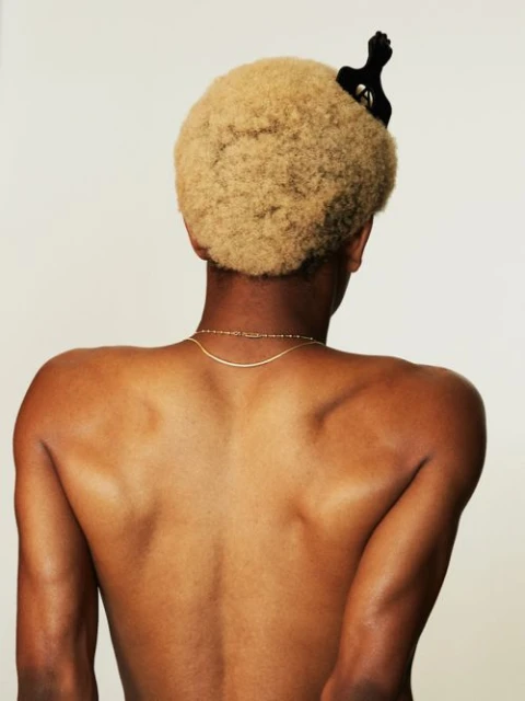 Close-up shot from behind of a shirtless person with blond afro-textured hair and a black hair clip against a white backdrop.