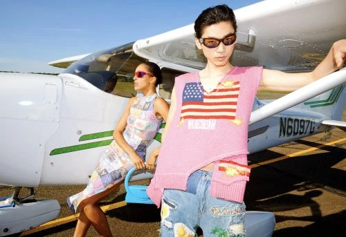 Two fashion models pose by a small airplane on a sunny day, wearing trendy clothing and sunglasses for a high-fashion editorial shot.