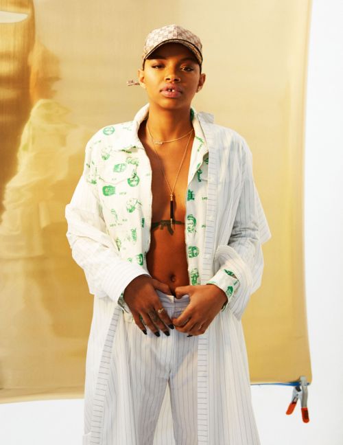 Young adult poses in a striped suit and patterned shirt, standing against a gold backdrop with a neutral expression and a cap.