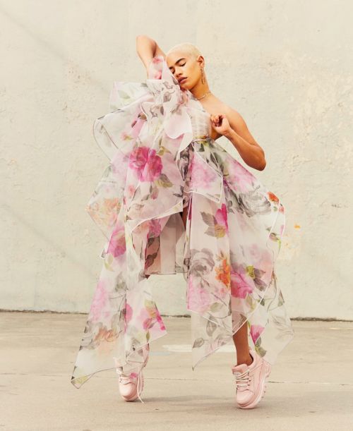A young adult posing outdoors in a large floral dress and pink platform shoes, with short buzz cut hair and gold jewelry.