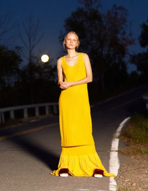 Woman in a flowing yellow dress standing on a road at night with a full moon, creating a striking fashion editorial shot.
