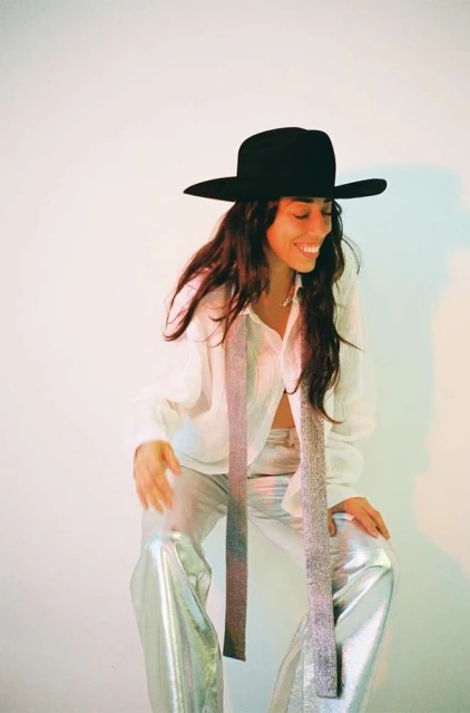 Woman in a wide-brimmed black hat and silver pants smiles in a studio setting, creating a chic and fashionable portrait.
