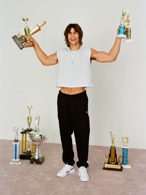 Smiling man stands surrounded by trophies in a photo studio, wearing a white shirt and black sweatpants, holding two trophies aloft.
