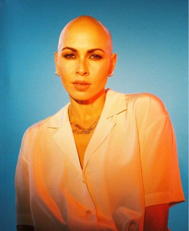 Portrait of a bald woman in a white satin button-down shirt against a blue backdrop under warm lighting, photographed by Jason Rodgers