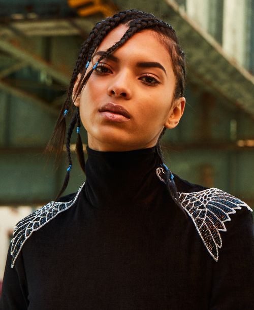 Close-up of a stylish adult female model with braided hair wearing a black turtleneck with ornate shoulder decoration and an industrial backdrop.