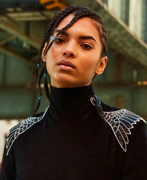 Close-up of a stylish adult female model with braided hair wearing a black turtleneck with ornate shoulder decoration and an industrial backdrop.