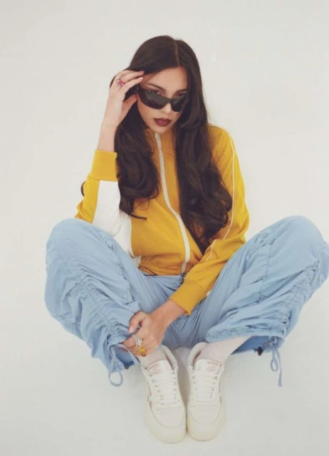 A woman with sunglasses and a tracksuit poses indoors with her legs crossed, featuring a bright yellow jacket and pale blue pants.