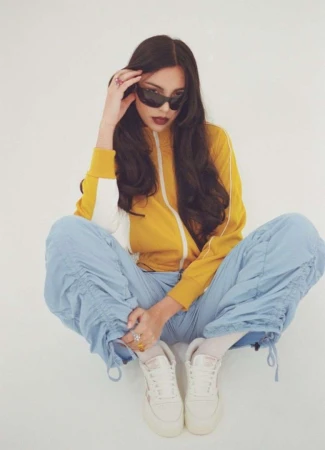 A woman with sunglasses and a tracksuit poses indoors with her legs crossed, featuring a bright yellow jacket and pale blue pants.