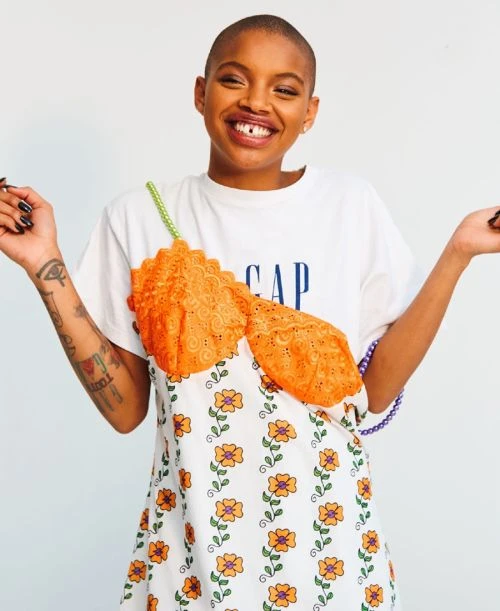 Smiling woman in white t-shirt dress with orange lace top and floral pattern. Posing in front of a white background with hands up.