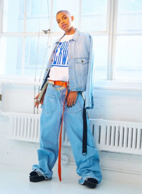 Young adult poses in light blue, wide-leg denim outfit and platform shoes against a white brick wall and bright windows, fashion shoot.