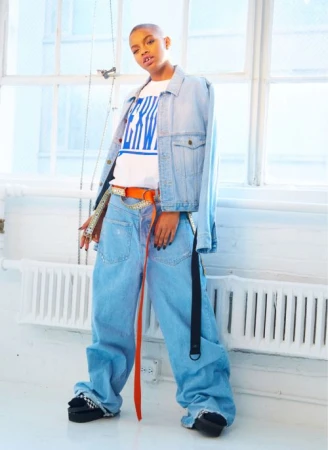 Young adult poses in light blue, wide-leg denim outfit and platform shoes against a white brick wall and bright windows, fashion shoot.