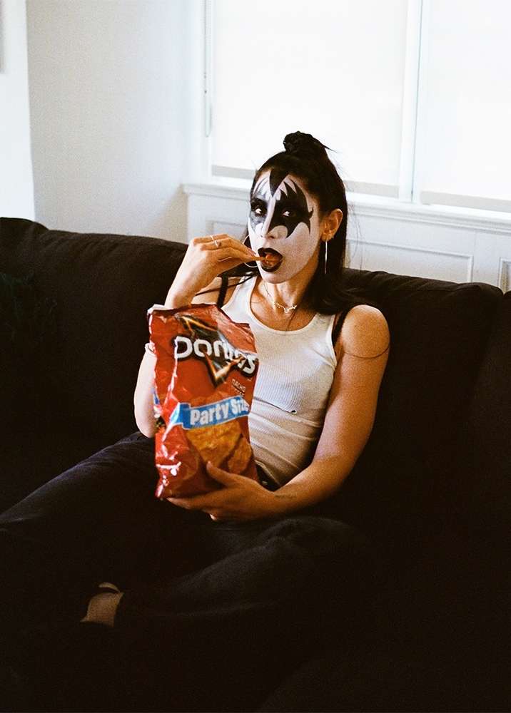 Woman in Kiss makeup sits on a couch eating Doritos. She has long dark hair and is wearing a white tank top and black pants in a casual setting.