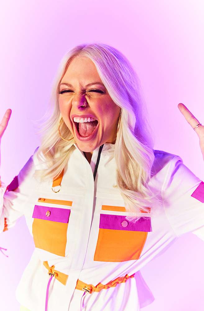 Excited woman with open mouth and raised hands against bright purple background. She wears a white jumpsuit with orange and pink pockets.