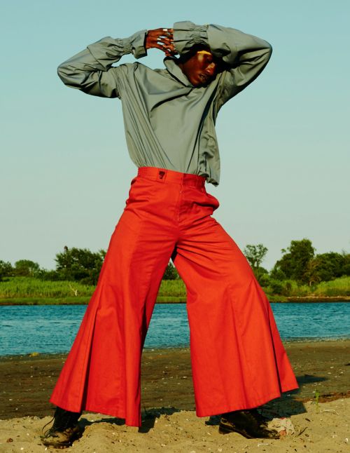 Fashionable African model poses on the beach in red flared pants and a draped gray shirt, with ocean and sky in the background.