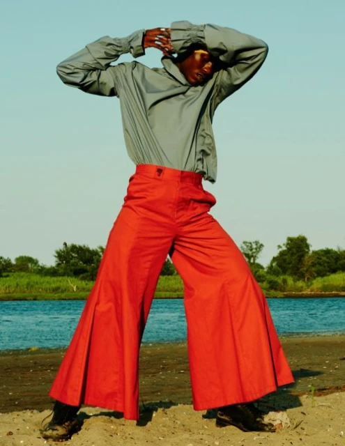 Fashionable African model poses on the beach in red flared pants and a draped gray shirt, with ocean and sky in the background.