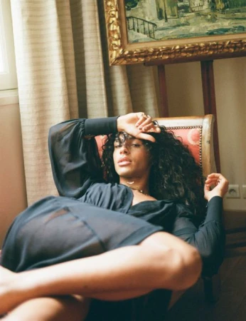 Woman with long, dark curly hair poses in a chair in a room with beige striped wallpaper, a framed painting, and white electrical outlets.