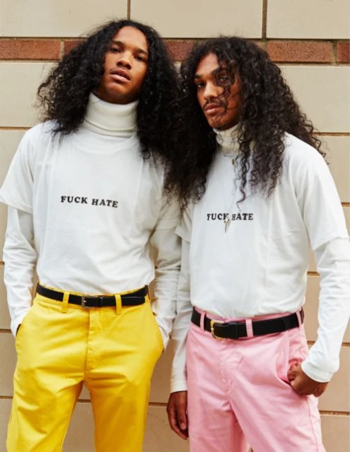 Two men with long curly hair pose wearing matching turtlenecks and t-shirts with the words "F* HATE" in front of a brick wall.