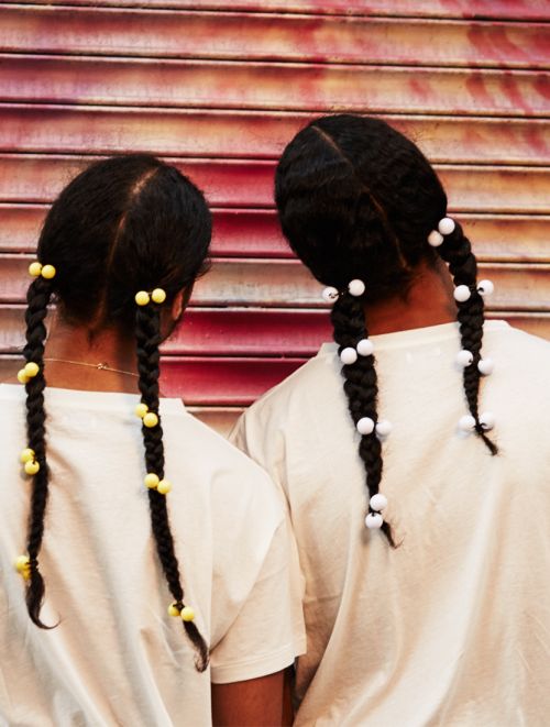 Two people stand close together showing their braided hair with yellow and white beads in front of a red and pink striped background.