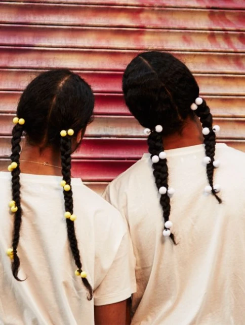 Two people stand close together showing their braided hair with yellow and white beads in front of a red and pink striped background.
