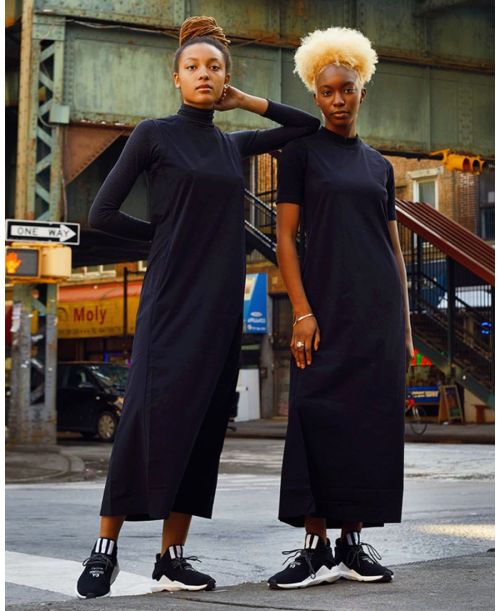 Two black women stand together in black dresses and sneakers, posing in a city street environment with buildings and traffic signals.