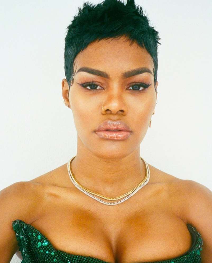 Portrait of a black woman with short dark hair, wearing a green sequined top and gold necklace, posing against a white backdrop.