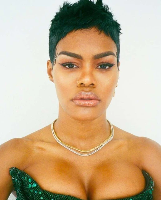 Portrait of a black woman with short dark hair, wearing a green sequined top and gold necklace, posing against a white backdrop.