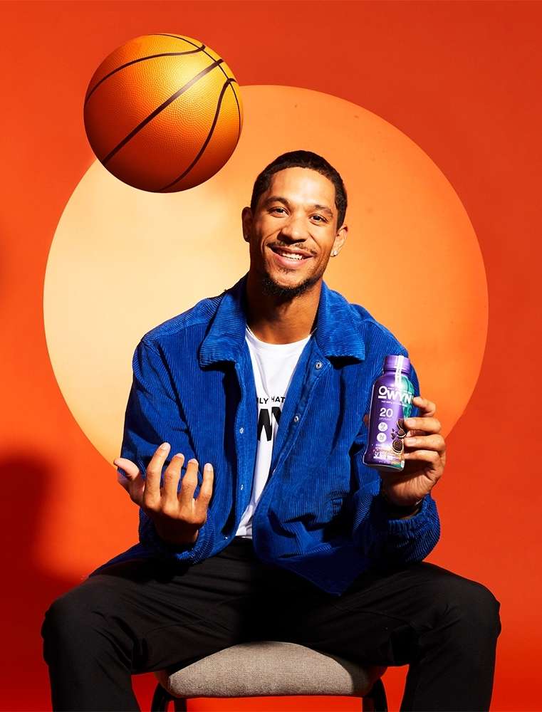 Man sitting on stool holding drink with basketball floating on orange background. He is wearing a blue corduroy jacket.