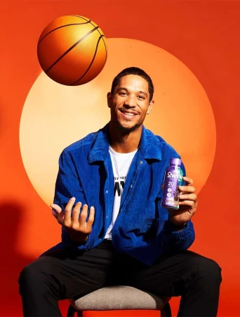 Man sitting on stool holding drink with basketball floating on orange background. He is wearing a blue corduroy jacket.