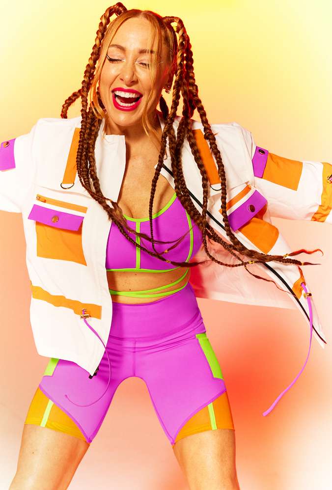 An energetic woman in athletic wear and braided hair dances against a warm-toned background, showcasing a vibrant and joyful expression.