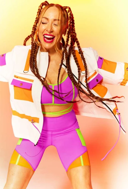 An energetic woman in athletic wear and braided hair dances against a warm-toned background, showcasing a vibrant and joyful expression.