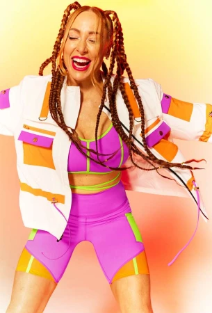 An energetic woman in athletic wear and braided hair dances against a warm-toned background, showcasing a vibrant and joyful expression.