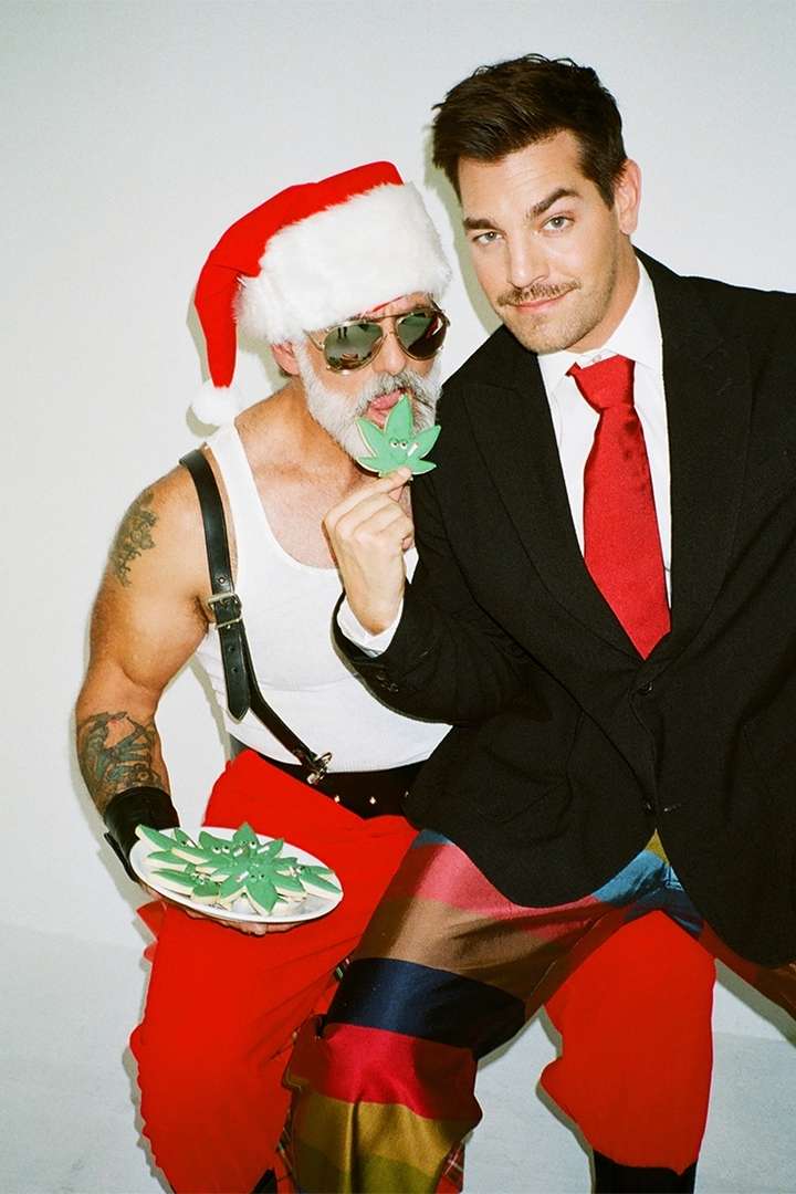 Two men pose in front of a white background: one dressed as Santa Claus, the other in a suit with colorful pants.  