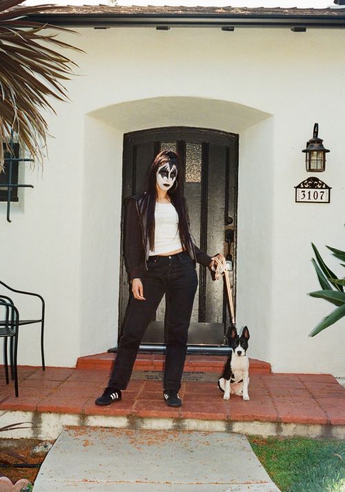 A person with theatrical makeup poses with a dog on a house porch, wearing a black jacket, jeans, and white top.