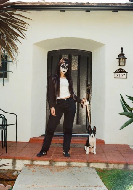 A person with theatrical makeup poses with a dog on a house porch, wearing a black jacket, jeans, and white top.
