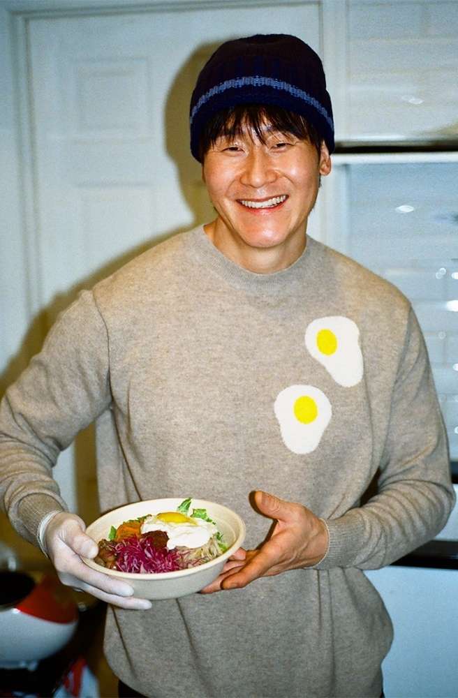 An Asian man smiles and holds a bowl of Korean bibimbap in a modern kitchen with white cabinets, wearing a sweater and hat.