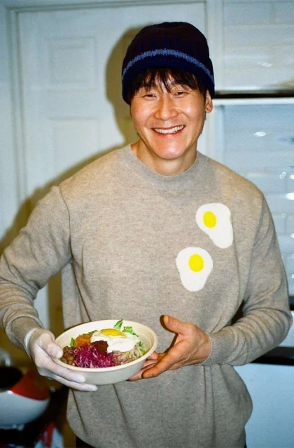 An Asian man smiles and holds a bowl of Korean bibimbap in a modern kitchen with white cabinets, wearing a sweater and hat.