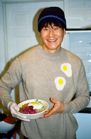 An Asian man smiles and holds a bowl of Korean bibimbap in a modern kitchen with white cabinets, wearing a sweater and hat.