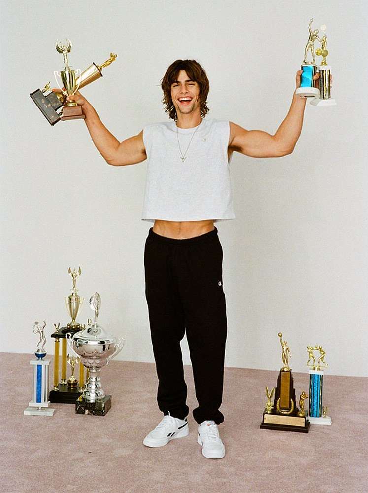 Man holding trophies in studio: A stylish male model in a crop top and black sweatpants poses with several gold and silver trophies.