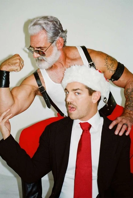 Two men pose indoors: an older man with grey hair and a beard flexes his arm, while a younger man wears a Santa hat and a suit.