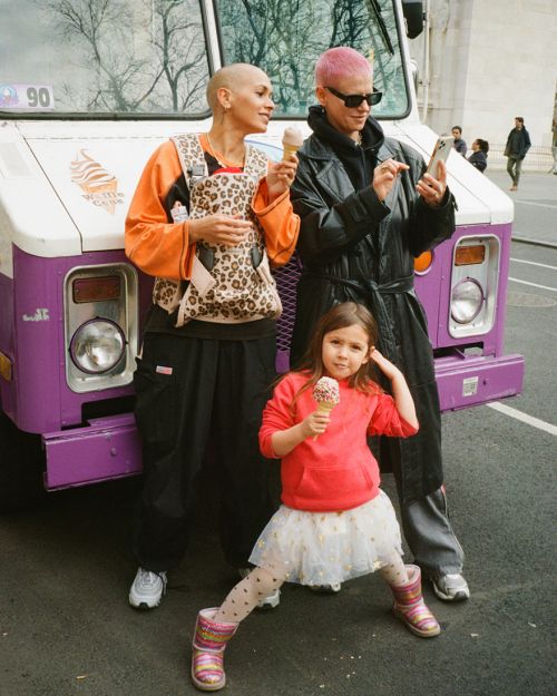 Two women and a child stand in front of a purple ice cream truck, enjoying ice cream cones on a city street during the daytime.