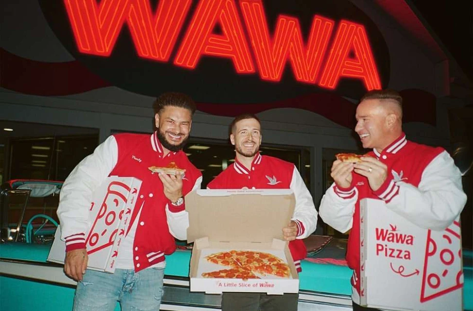 Three adult men in red letterman jackets stand outdoors, eating pizza from Wawa Pizza boxes in front of a Wawa store at night.