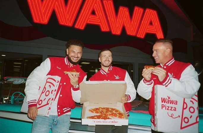 Three adult men in red letterman jackets stand outdoors, eating pizza from Wawa Pizza boxes in front of a Wawa store at night.