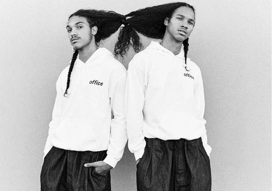 Black and white shot of two adult men with long curly braided hair wearing matching white hoodies standing close together against a neutral wall.