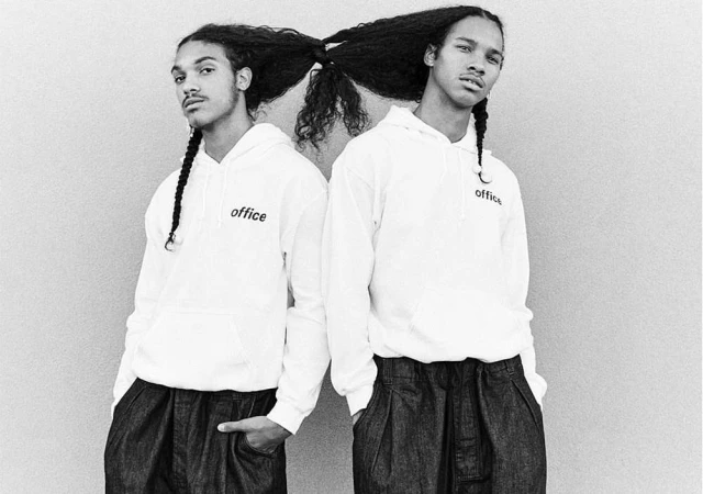 Black and white shot of two adult men with long curly braided hair wearing matching white hoodies standing close together against a neutral wall.