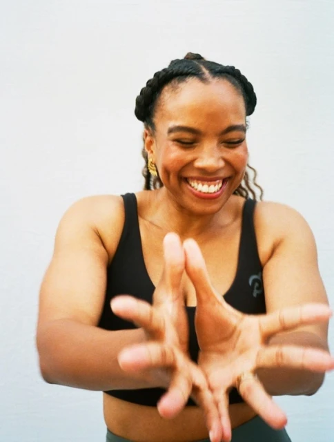 Smiling African-American woman in activewear poses indoors with outstretched hands against a bright white background in a medium shot.
