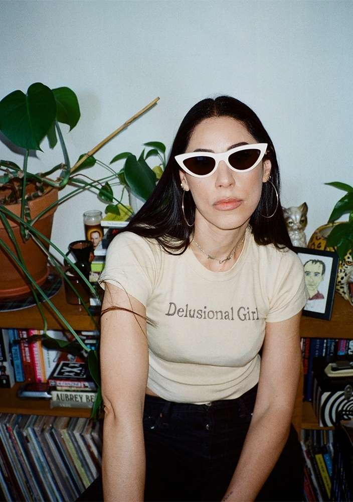 Young woman in sunglasses and a t-shirt sitting indoors near plants and books on shelves looking at the camera in medium shot.