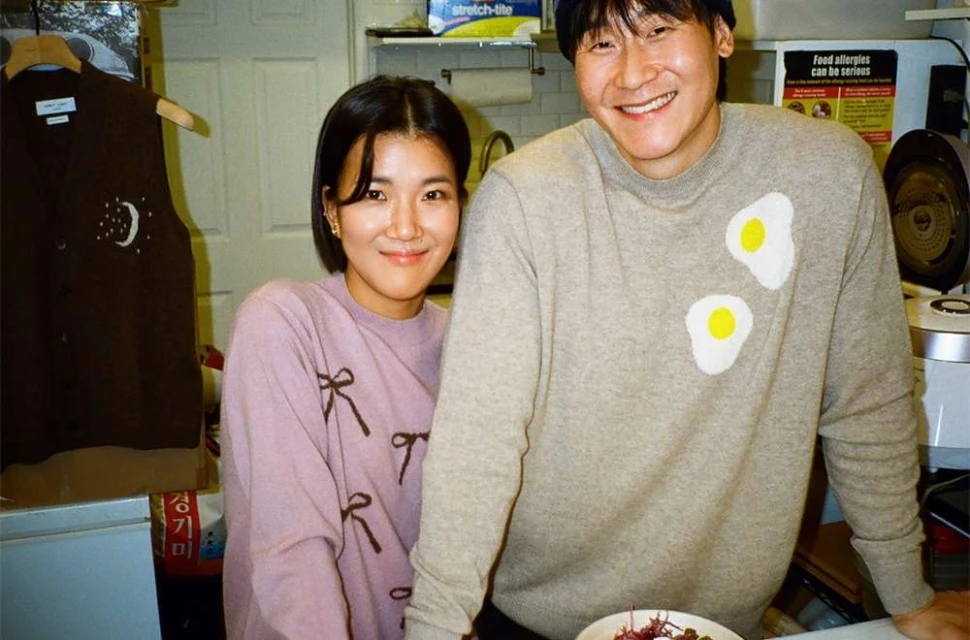 Smiling Asian couple poses indoors. Woman wears pink bow-patterned sweater; man wears light brown sweater with fried egg design.
