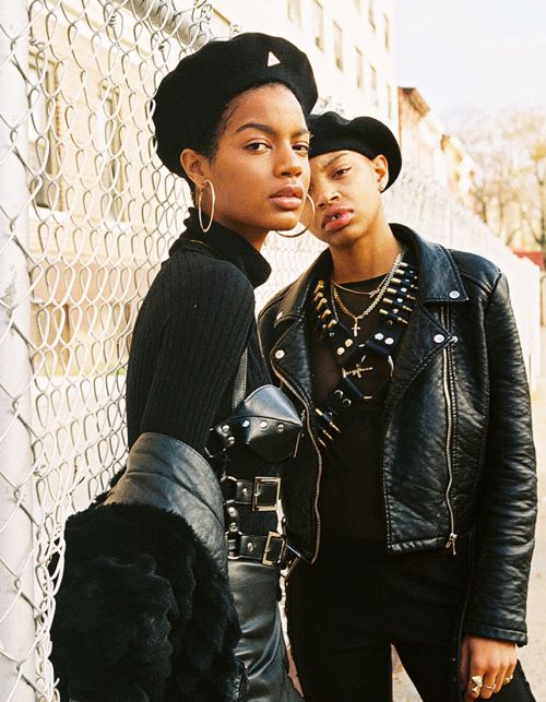 Two young women pose in berets and leather jackets against a chain-link fence and brick building in a medium outdoor shot.