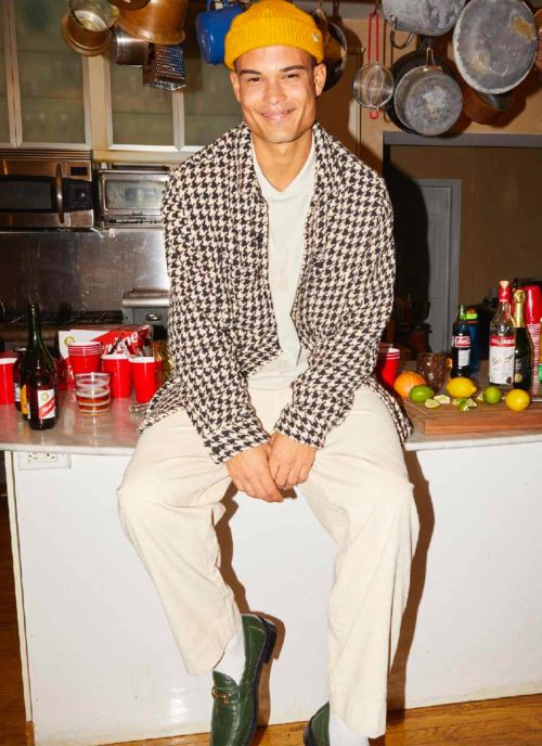 A smiling man wearing a houndstooth jacket and yellow beanie sits on a counter in a kitchen with drink and glassware.